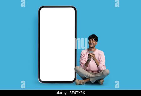 Happy indian guy hipster sitting by big smartphone, mockup Stock Photo