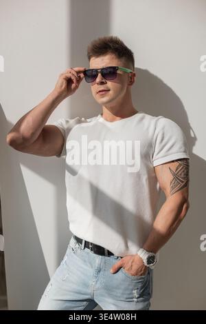 Young fit man with arm tattoos in casual outfit with t-shirt. Wearing sunglasses, casual white tee and jeans in a trendy, bright apartment. Stock Photo