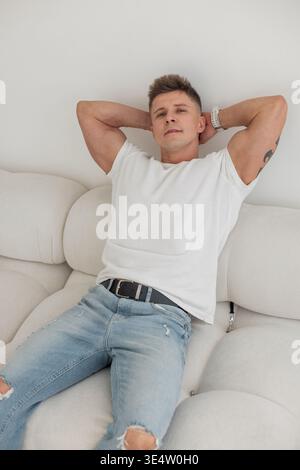 Young man in ripped jeans and t-shirt resting comfortably on a soft couch. Minimalist lifestyle and home comfort concept. Stock Photo
