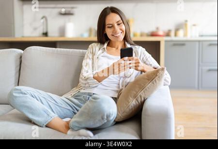 Cheerful young woman texting on her smartphone, sitting cross-legged on comfortable couch Stock Photo