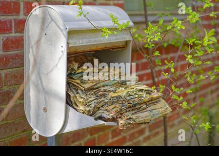 Rusty metal mailbox attached to brick wall filled with old weathered newspapers showing neglect and exposure to elements outdoors. Stock Photo