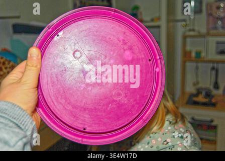 A hand holds a scratched pink plastic lid indoors with a blurred person and household items in the background, showing everyday life and casual settin Stock Photo