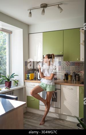 Cheerful woman standing in kitchen looking at window and enjoying morning coffee on weekend. Stock Photo