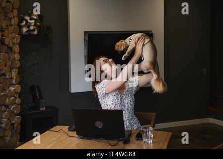 Joyful woman lifting cute dog while working on laptop at home. Authentic moment of remote work lifestyle, pet companionship and taking a break from Stock Photo