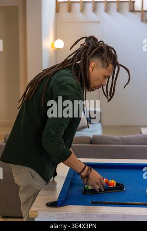 African American man wearing dark green jacket leaning on blue pool table arranging rack of balls Stock Photo