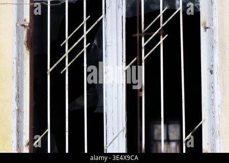 Dilapidated window frame with security bars and shattered glass panes, showing decay and neglect behind dark interior Stock Photo