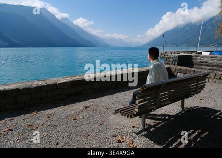 man sitting on a wooden bench by Lake Brienz, Switzerland, looking at the calm water and mountain ranges Stock Photo
