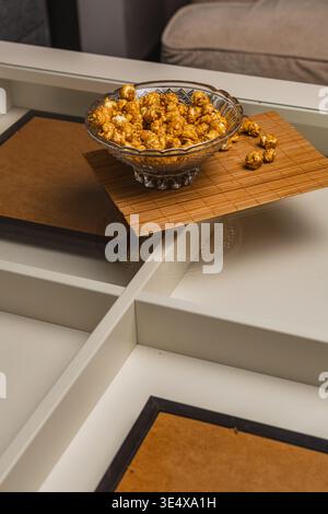 A bowl filled with popcorn sits on a brown mat placed on a white table. A soft couch can be seen in the background. It looks like a moment for snacks. Stock Photo