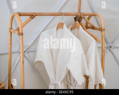 Couple white waffle bathrobes hanging on wooden rack in modern white room, representing spa luxury, hotel and resort amenities, relaxation, wellness l Stock Photo