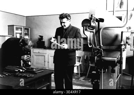 Sloviansk, 1950s-1960s, Armature-Insulator Plant (AIZ). Two technical specialists perform rigorous quality control on heavy metal components inside a bright factory laboratory. One engineer stands examining a part, while his seated colleague uses a magnifying instrument to inspect a batch of manufactured hardware on the wooden desk. A large industrial testing machine stands nearby. This historical scene highlights meticulous engineering labor, preserving the authentic atmosphere of life before the war and the industrial heritage of a peaceful Donbas. Stock Photo
