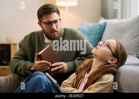 Caucasian middle aged man holding book talking to Caucasian preteen girl wearing glasses sitting on floor leaning against sofa listening with thoughtful expression Stock Photo