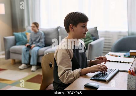 Caucasian boy typing on computer keyboard at desk with notebook open, sitting in modern living room, another Caucasian child in background playing video game on couch, both focused on activities Stock Photo