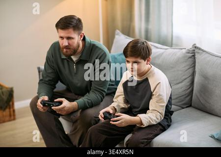 Caucasian middle aged man and Caucasian boy sitting on sofa playing video game together, holding controllers focused on screen spending leisure time in living room Stock Photo