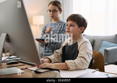 Caucasian boy sitting at desk using desktop computer while Caucasian girl standing beside holding digital tablet, both children focusing on screen in home environment Stock Photo