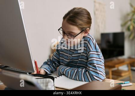 Caucasian girl child wearing glasses sitting at desk writing in notebook while looking at computer monitor, studying or doing homework in home environment with focus and concentration Stock Photo