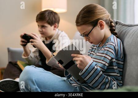 Caucasian girl with glasses sitting on sofa using digital tablet while Caucasian boy sitting beside playing video game with controller, both appearing focused and engaged in activities Stock Photo