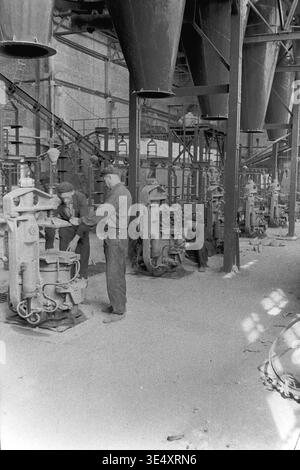 Sloviansk, 1950s-1960s, Armature-Insulator Plant (AIZ). A wide view of the industrial factory floor featuring massive overhead metal hoppers and a row of mechanical forming presses. Several male workers are actively operating the heavy machinery to manufacture ceramic components. This highly detailed historical archive photograph perfectly illustrates the complex production line infrastructure, organized physical labor, and the impressive scale of Soviet manufacturing during the era of a peaceful Donbas. Stock Photo