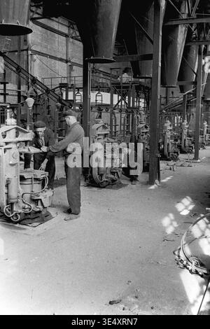 Sloviansk, 1950s-1960s, Armature-Insulator Plant (AIZ). A wide view of the industrial factory floor featuring massive overhead metal hoppers and a row of mechanical forming presses. Several male workers are actively operating the heavy machinery to manufacture ceramic components. This highly detailed historical archive photograph perfectly illustrates the complex production line infrastructure, organized physical labor, and the impressive scale of Soviet manufacturing during the era of a peaceful Donbas. Stock Photo