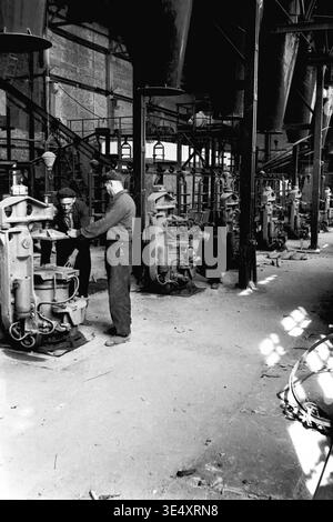 Sloviansk, 1950s-1960s, Armature-Insulator Plant (AIZ). A wide view of the industrial factory floor featuring massive overhead metal hoppers and a row of mechanical forming presses. Several male workers are actively operating the heavy machinery to manufacture ceramic components. This highly detailed historical archive photograph perfectly illustrates the complex production line infrastructure, organized physical labor, and the impressive scale of Soviet manufacturing during the era of a peaceful Donbas. Stock Photo