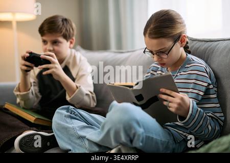 Caucasian boy wearing glasses reading book while sitting on sofa next to another Caucasian boy playing video game with controller, both children focused on separate activities indoors Stock Photo