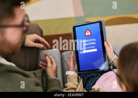Caucasian child holding tablet displaying parental control password screen while sitting next to Caucasian adult, both interacting on sofa, adult hand resting near child, close up view Stock Photo