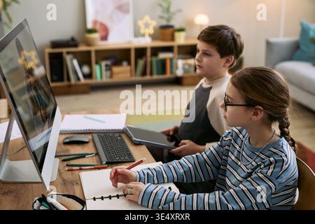 Caucasian girl and Caucasian boy sitting at desk using desktop computer for studying, girl writing in notebook while boy holding digital tablet, both focused on screen Stock Photo