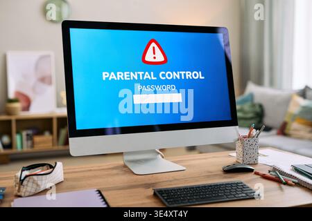Modern desktop computer displaying parental control password warning on screen, keyboard and office supplies scattered on wooden desk, home interior blurred in background Stock Photo