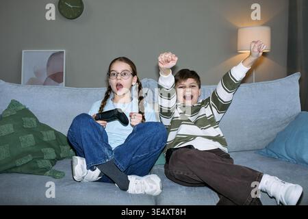 Caucasian girl with glasses holding game controller sitting next to Caucasian boy cheering with raised arms on sofa both children, appearing excited while playing video game together Stock Photo