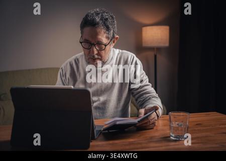 Middle aged man analyzing financial notes and reviewing data on tablet at home during evening. Concept of personal finance management, budgeting, inde Stock Photo