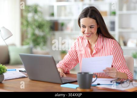 Happy freelance worker is holding form and using laptop working at home Stock Photo