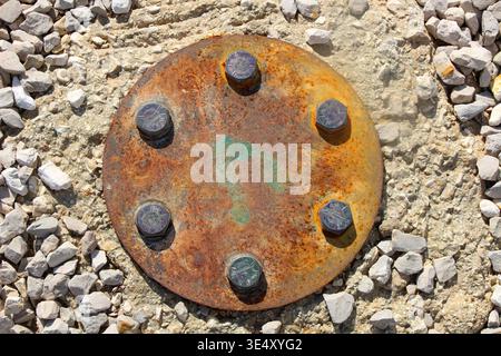 Rusty circular metal plate with six bolts securing a structural anchor, set into concrete and surrounded by loose gravel, representing industrial cons Stock Photo