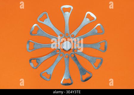 Twelve bottle openers are arranged in a clock shape around a metal crown cap on an orange background Stock Photo