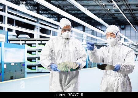 Team of engineers doing research and development on silicon wafer, brainstorming in cleanroom. Teamworking colleagues in protective suit examining microelectronic semiconductor substrate for defects Stock Photo
