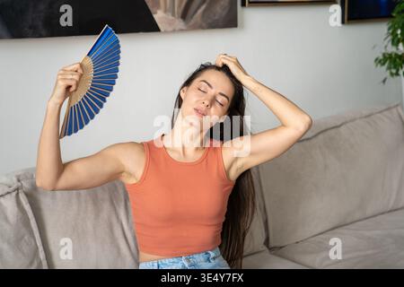 Glad woman successfully coping with abnormal heat, blowing with paper fan, sitting on couch. Stock Photo