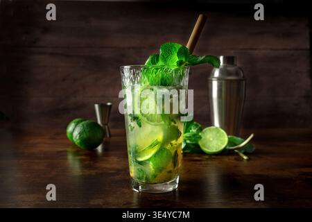 Mojito, fresh summer cocktail with mint, lime, and ice, side view of a single glass on a wooden table, with a shaker Stock Photo