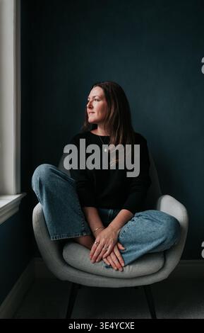 Attractive middle aged woman sitting in chair beside window. Stock Photo