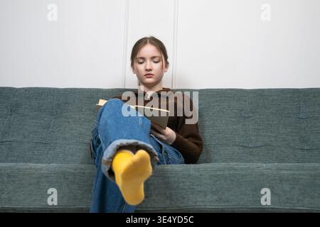 Teen girl sitting comfortably on a sofa at home, engrossed in reading a book, representing concepts of learning, leisure, education, and peaceful rela Stock Photo