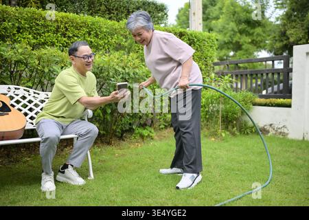 Senior couple enjoying gardening together in their home garden. A woman watering plants with a hose while her husband relaxes on a bench using a smartphone, showing a happy retirement lifestyle || Model released Stock Photo