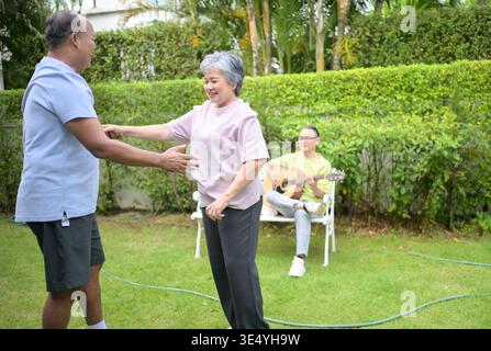 Group of asian seniors singing and dancing while their friend plays guitar in a garden, enjoying friendship and outdoor leisure activity || Model released Stock Photo