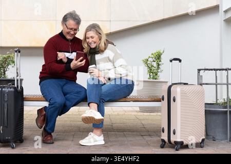 Senior couple sitting on bench by wall, sharing phone while holding coffee cup with two suitcases. Luggage, smartphone, beverage, companionship, outdoor, urban, planters || Model released Stock Photo