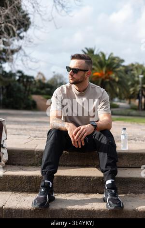 Stylish traveler hipster man with vintage sunglasses in fashion outfit sits on steps and relaxes on mountain in Barcelona Stock Photo