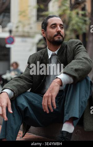 Fashion handsome hipster man in trendy casual clothes with coat sitting on street in Barcelona Stock Photo