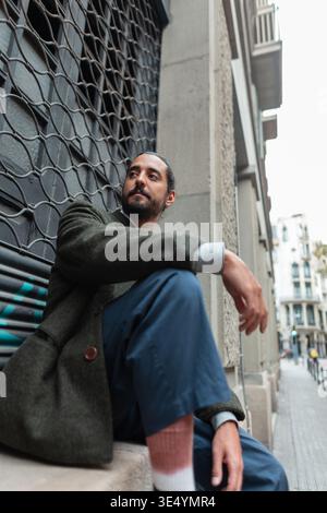 Urban fashion handsome man in stylish clothes with coat and pants sitting and posing on street in Barcelona city Stock Photo