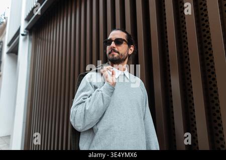 Handsome fashion man with beard and moustache with sunglasses in fashionable casual outfit walking in the city near bronze metal wall Stock Photo