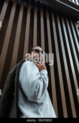 Stylish urban man with sunglasses in fashion casual clothes with coat walks in the city near bronze metal wall Stock Photo