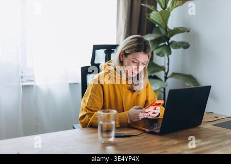 Woman distracted by smartphone while working on laptop in home office showing multitasking and digital interruption in remote work Stock Photo