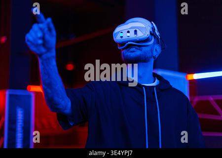 Happy handsome man in vr glasses playing and trying to touch something when exploring simulation of world in the dark with neon light. Technology and Stock Photo