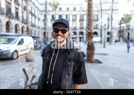 Happy fashion beautiful hipster man with cool sunglasses in black cap with denim jacket and hoodie walking in Barcelona Stock Photo