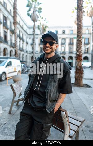 Happy handsome fashion man with smile with sunglasses with black cap in black denim clothes with hoodie stands near bench in Barcelona city Stock Photo