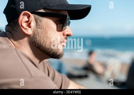 Handsome man with beard with fashion sunglasses with black cap in beige T-shirt sits on beach on seashore. Stock Photo